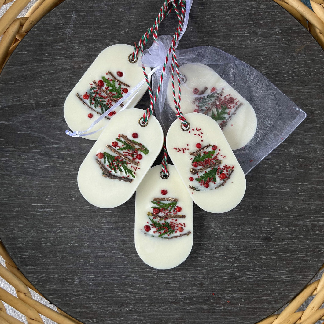 An elongated oval white wax tablet embellished with tiny dried branches, cranberry seeds, and red peppercorns resembles a Christmas tree. It features a red and green decorative rope threaded through a metal eyelet. Packaging: white organza bag.