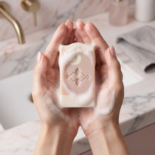Load image into Gallery viewer, Pink and cream collagen soap bar with embossed logo held in foamy hands.