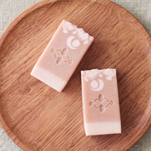 Load image into Gallery viewer, Two collagen soap bars in flat lay, blush-peach with cream base, white floral accents, stamped emblem, and handmade texture