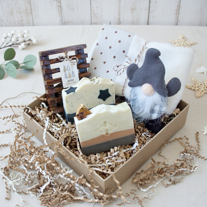 Gift box featuring two handmade artisan soaps with decorative designs, a rustic wooden soap dish with a “Merry Christmas” tag, a soft white and brown polka dot towel, and a small Nordic-style gnome doll, all nestled in eco-friendly shredded paper.