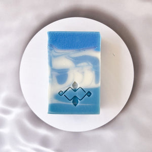 A square soap bar with vivid blue and white swirls features a stamped Woods and Mosses logo on its smooth surface. The design evokes a sense of Arctic freshness, with striking contrasts between the rich blue and crisp white colors.