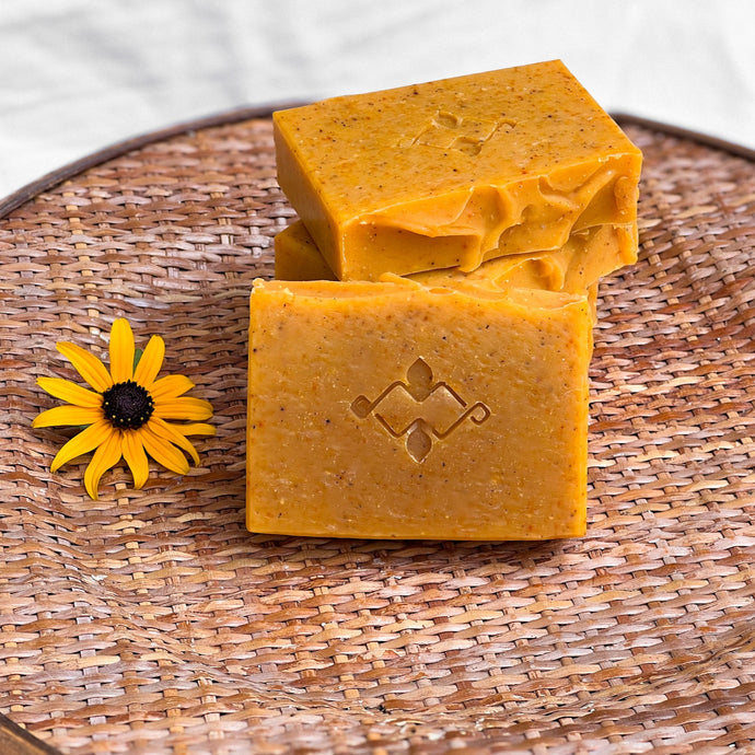 An orange square soap with small brownish dots throughout. The bar has a slightly wavy top and a company logo stamped in the center.