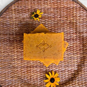 An orange square soap with small brownish dots throughout. The bar has a slightly wavy top and a company logo stamped in the center.