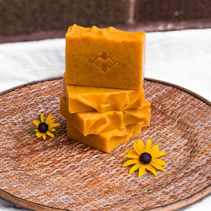 An orange square soap with small brownish dots throughout. The bar has a slightly wavy top and a company logo stamped in the center.