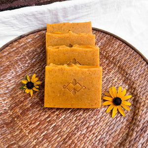 An orange square soap with small brownish dots throughout. The bar has a slightly wavy top and a company logo stamped in the center.