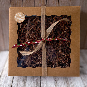 Men's Self Care Gift Basket for Dad - Natural Skincare Christmas Set: A brown kraft gift box with a clear window is filled with raffia paper and tied with brown and dark red ribbons. It features a Merry Christmas decorative sticker.