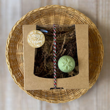 Load image into Gallery viewer, A kraft box with a transparent lid filled with brownish soap, tied with a festive white, red, and green rope and adorned with a Merry Christmas sticker. A round pale green mini soap featuring a delicate snowflake print on the top.