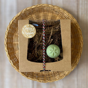 A kraft box with a transparent lid filled with brownish soap, tied with a festive white, red, and green rope and adorned with a Merry Christmas sticker. A round pale green mini soap featuring a delicate snowflake print on the top.