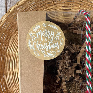 A kraft box with a transparent lid filled with brownish soap, tied with a festive white, red, and green rope and adorned with a Merry Christmas sticker.