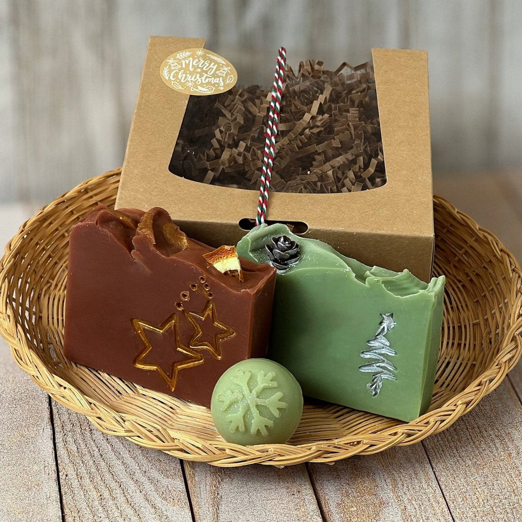 A kraft box with a transparent lid filled with brownish soap, tied with a festive white, red, and green rope and adorned with a Merry Christmas sticker. Beside the box are brownish square soap, green square soap, and a round pale green mini soap.