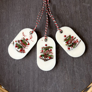 An elongated oval white wax tablet embellished with tiny dried branches, cranberry seeds, and red peppercorns resembles a Christmas tree. It features a red and green decorative rope threaded through a metal eyelet.