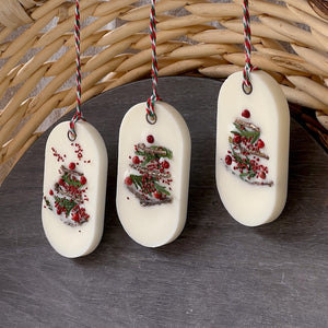 An elongated oval white wax tablet embellished with tiny dried branches, cranberry seeds, and red peppercorns resembles a Christmas tree. It features a red and green decorative rope threaded through a metal eyelet.