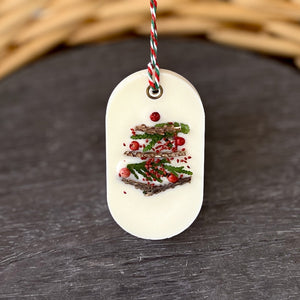 An elongated oval white wax tablet embellished with tiny dried branches, cranberry seeds, and red peppercorns resembles a Christmas tree. It features a red and green decorative rope threaded through a metal eyelet.