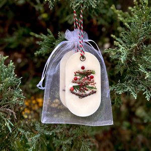 An elongated oval white wax tablet embellished with tiny dried branches, cranberry seeds, and red peppercorns resembles a Christmas tree. It features a red and green decorative rope threaded through a metal eyelet. Packaging: white organza bag.