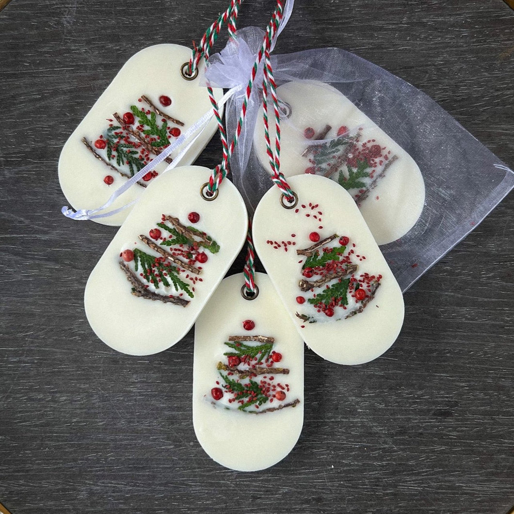 An elongated oval white wax tablet embellished with tiny dried branches, cranberry seeds, and red peppercorns resembles a Christmas tree. It features a red and green decorative rope threaded through a metal eyelet. Packaging: white organza bag.