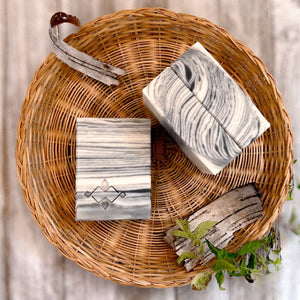 A square soap bar with a birch bark pattern in white, gray, and black, featuring a stamped Woods and Mosses logo in the corner. Ideal for gifting or as a decorative bathroom accent.