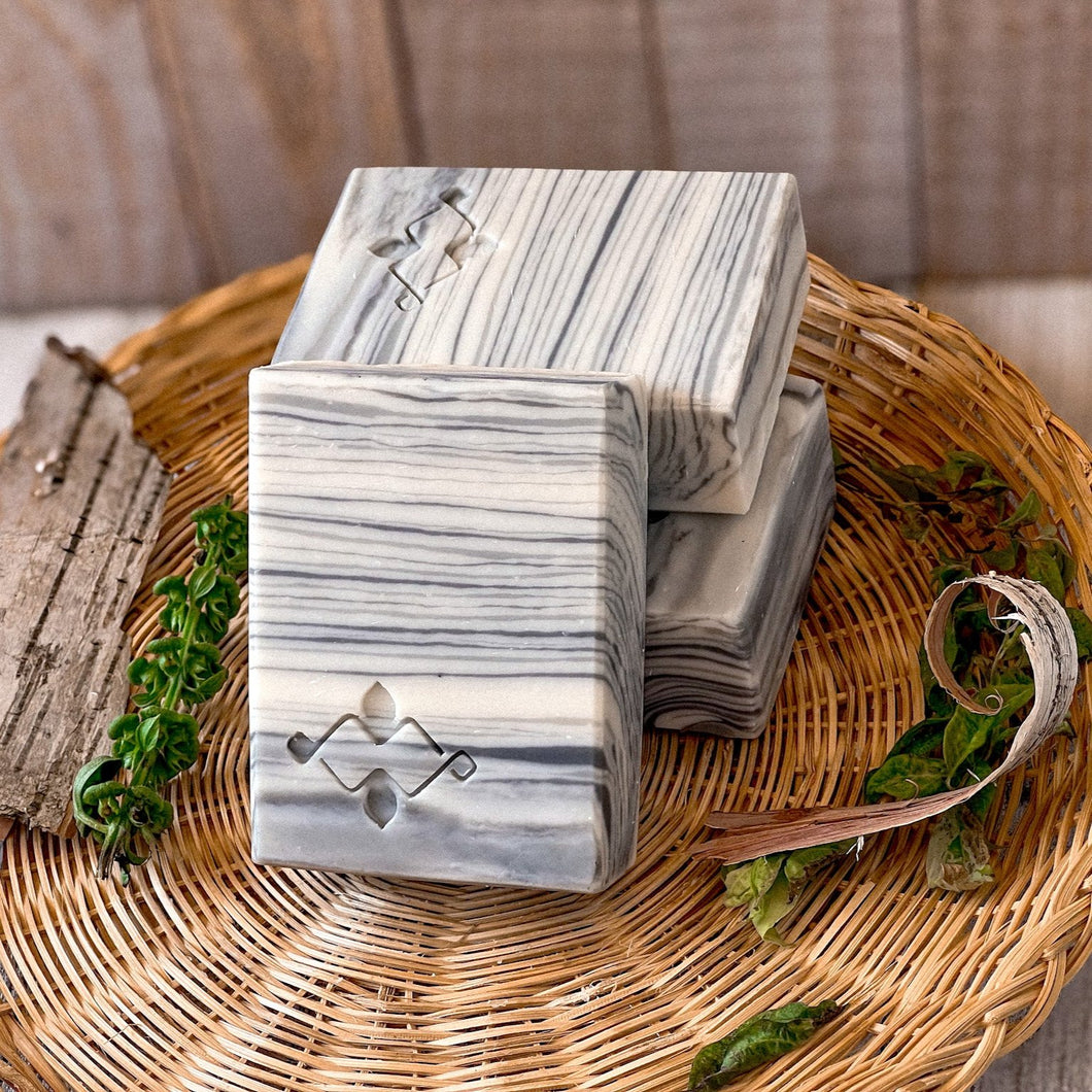 A square soap bar with a birch bark pattern in white, gray, and black, featuring a stamped Woods and Mosses logo in the corner. Ideal for gifting or as a decorative bathroom accent.