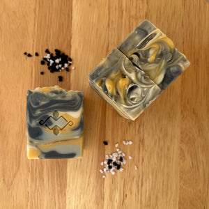 A square, tall, layered soap with distinct white, black, and yellow layers, featuring a textured top. The center showcases a Woods and Mosses company logo, adding a unique touch to the soap’s design.