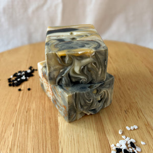 A square, tall, layered soap with distinct white, black, and yellow layers, featuring a textured top. 