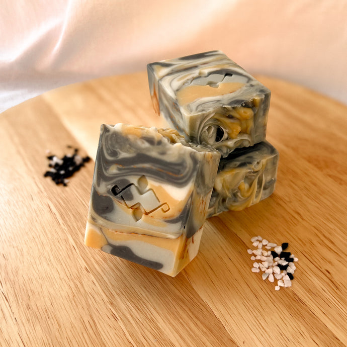 A square, tall, layered soap with distinct white, black, and yellow layers, featuring a textured top. The center showcases a Woods and Mosses company logo, adding a unique touch to the soap’s design.