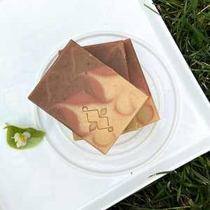 Handmade soap bar with beige, dusty rose, and brown swirls. The Woods and Mosses company logo is prominently displayed in the lower corner.