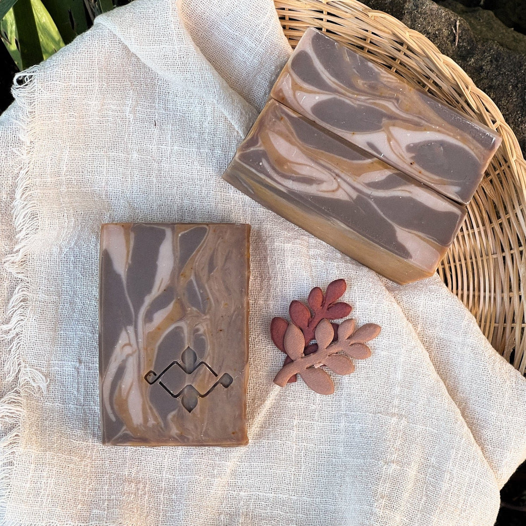 Two handcrafted soap bars with a marbled caramel and chocolate design are placed on a beige cloth. One bar features a stamped Woods and Mosses logo while the other showcases smooth swirls.