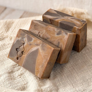 3 handcrafted, rectangular soap bars with a marbled caramel and chocolate design, sitting on a textured beige fabric. The soap bars have a smooth, swirled pattern with a Woods and Mosses stamped logo on the surface, giving them an artisanal look.