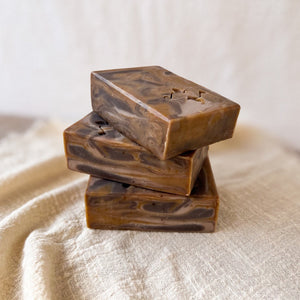 3 handcrafted, rectangular soap bars with a marbled caramel and chocolate design, sitting on a textured beige fabric. The soap bars have a smooth, swirled pattern with a Woods and Mosses stamped logo on the surface, giving them an artisanal look.