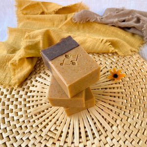 A handcrafted soap bar with two distinct layers: a light beige base and a brown top, divided by a visible seam. The soap has a decorative stamp on the front.