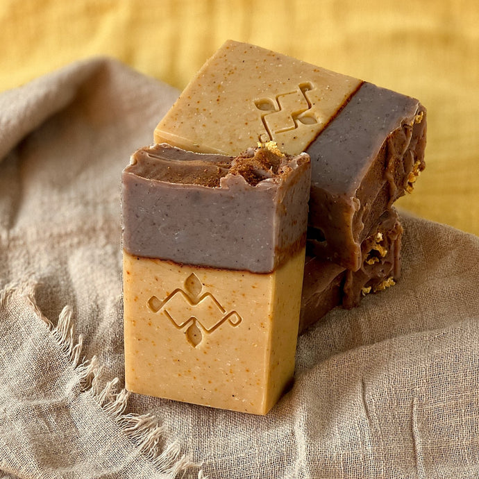 A handcrafted soap bar with two distinct layers: a light beige base and a brown top, divided by a visible seam. The soap has a Woods and Mosses company stamp on the front.