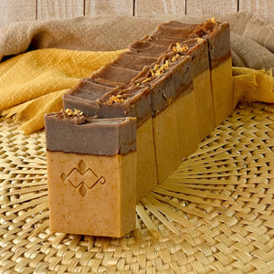 A row of handcrafted soap bars with a light beige base and dark brown tops, featuring a Woods and Mosses company stamp on the front. The soap tops are textured, adorned with orange peel.