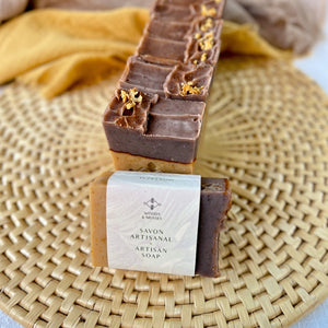 A row of handcrafted soap bars with a beige base and brown tops, adorned with orange peel. In front of the row is a single soap wrapped with a label from Woods & Mosses that reads Savon Artisanal and Artisan Soap.