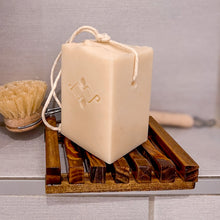 Load image into Gallery viewer, A square, creamy-colored soap features a sturdy rope threaded through a corner hole for easy hanging and drying. It is placed on a brown wooden drained soap dish. In the back: round-headed brush with natural bristles and a wooden handle.