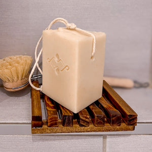 A square, creamy-colored soap features a sturdy rope threaded through a corner hole for easy hanging and drying. It is placed on a brown wooden drained soap dish. In the back: round-headed brush with natural bristles and a wooden handle.