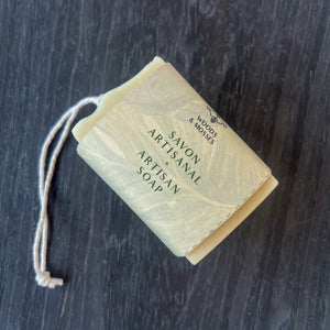 A rectangular soap bar with a simple rope for hanging, wrapped in light green paper with Savon Artisanal / Artisan Soap and Woods & Mosses branding in a minimalist font.