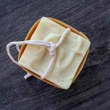 Load image into Gallery viewer, A square, creamy-colored soap bar with a natural, minimalist appearance. It features a sturdy rope threaded through for easy hanging and drying. The soap has a textured top.