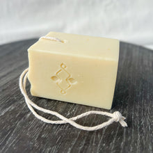 Load image into Gallery viewer, A square, creamy-colored soap bar with a natural, minimalist appearance. It features a sturdy rope threaded through a corner hole for easy hanging and drying. The soap has a stamped company logo on one side and a textured top.