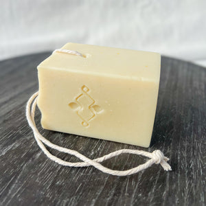 A square, creamy-colored soap bar with a natural, minimalist appearance. It features a sturdy rope threaded through a corner hole for easy hanging and drying. The soap has a stamped company logo on one side and a textured top.