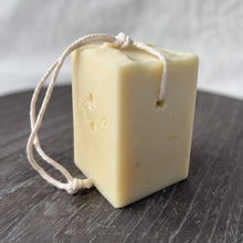 Load image into Gallery viewer, A square, creamy-colored soap bar with a natural, minimalist appearance. It features a sturdy rope threaded through a corner hole for easy hanging and drying. The soap has a stamped company logo on one side and a textured top.