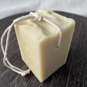 A square, creamy-colored soap bar with a natural, minimalist appearance. It features a sturdy rope threaded through a corner hole for easy hanging and drying. The soap has a stamped company logo on one side and a textured top.