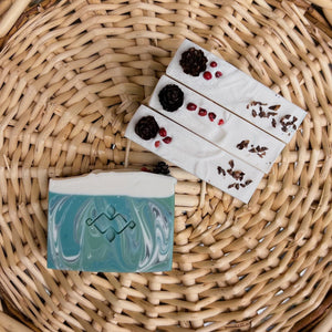 Textured, square soap bar swirled with green, white, and blue hues. Features a central Woods and Mosses company logo and a glitter-dusted pine cone on top.