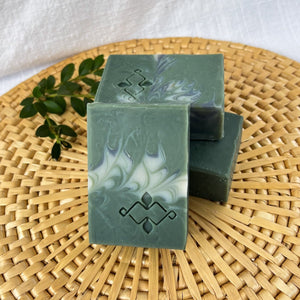 Square soap bars in dark and light sage green, with grey and white swirls and a stamped Woods and Mosses company logo on the surface.