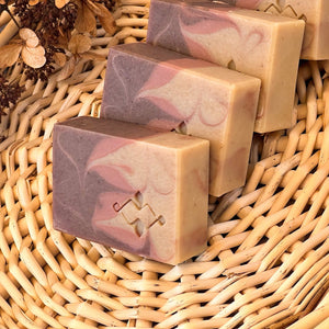 Handmade soap bar with beige, dusty rose, and brown swirls. The Woods and Mosses company logo is prominently displayed in the lower corner.