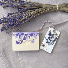 Load image into Gallery viewer, Luxury Lavender Spa Gift Basket, Christmas Edition by Woods and Mosses: A square soap bar is white with purple swirls and features a company logo. A rectangular white wax tablet is embellished with lavender and heather dried flowers. It features a white and beige decorative rope threaded through a metal eyelet.