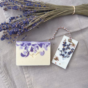 Luxury Lavender Spa Gift Basket, Christmas Edition by Woods and Mosses: A square soap bar is white with purple swirls and features a company logo. A rectangular white wax tablet is embellished with lavender and heather dried flowers. It features a white and beige decorative rope threaded through a metal eyelet.