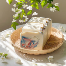 Load image into Gallery viewer, White jasmine soap with colorful design on a woven tray with flowers in the background - Woods and Mosses