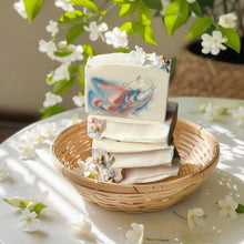 Load image into Gallery viewer, Stack of swirled jasmine soap bars in a woven basket with white flowers and green leaves in the background