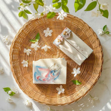 Load image into Gallery viewer, Three soap bars with decorative designs on a woven plate surrounded by white flowers and green leaves.