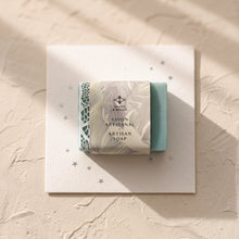 Load image into Gallery viewer, Greenish-blue artisan soap partly wrapped in a Woods &amp; Mosses paper sleeve, with white lace detail visible.