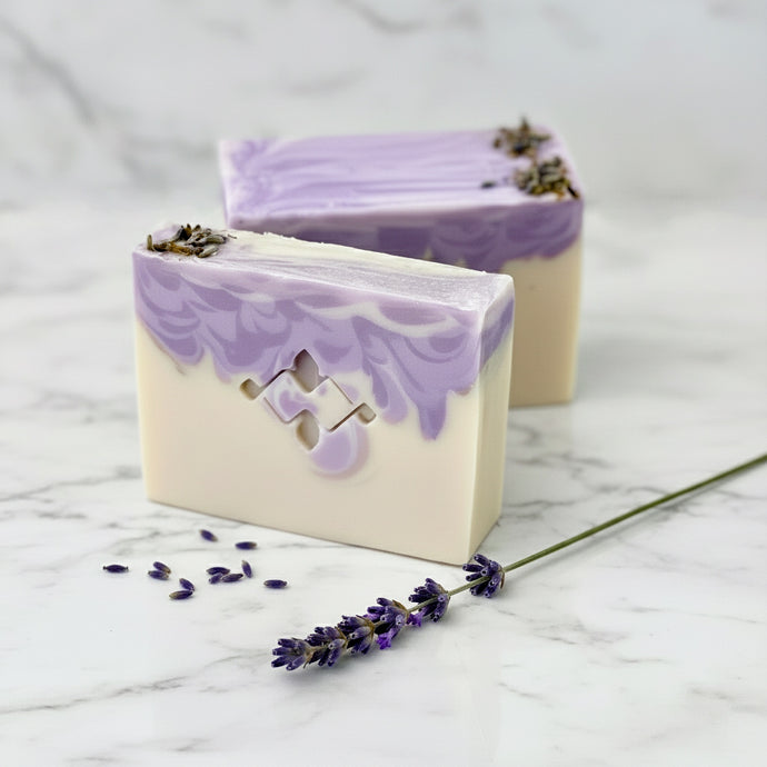 Handmade lavender soap bars featuring a smooth ivory base and a vibrant purple marbled top. One bar reveals Woods and Mosses logo, both are garnished with dried lavender buds for a natural touch.